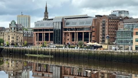 Newcastle Crown Court reflected in the River Tyne running in front of it. It is an imposing building made from smooth red stone with massive black windows and tall columns along its frontage.