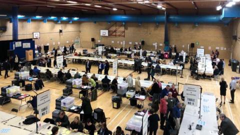 A sports hall at a leisure centre where votes are being counted after the elections in Reigate and Banstead in 2024. There are white tables with people sitting at them, doing the counting. There are also signs indicating the names of the different wards.