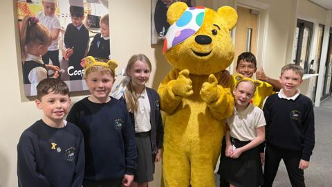 Pudsey bear - a yellow mascot with a colourful band over one eye - posing with children at a primary school. The children are all dressed in navy blue uniforms. Two of them have Pudsey ears on their heads. They are all looking at the camera and smiling.