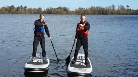 Two men wearing black clothing and life jackets are on stand-up paddleboards on a body of water, with trees in the distance behind them.
