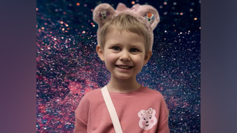 A little girl wearing a pink top and pink bear ears on top of her head stands smiling at the camera. She has short brown, blonde hair and a pink bear logo on her top.