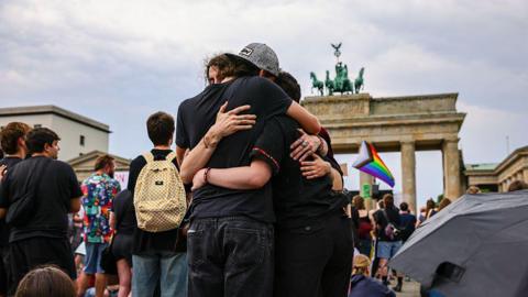 Three people hug each other with a rainbow LGBT flag visible in the background and the Brandenburg Gate behind