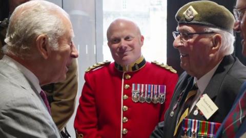 King Charles III meets veteran Richard Brock, who is wearing a green beret and his medals. Other members of the armed forces look on as the two speak. 
