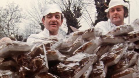 Two men in baker's uniforms stood behind a huge pile of hot cross buns on a market stall. 