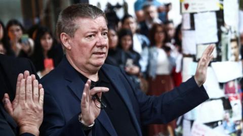 Liam Payne's father, Geoff, stands with his hands out acknowledging fans, not pictured, outside the hotel in Argentina where his son died. Geoff Payne is pictured side on, looking to the right and wearing a black polo shirt with a navy blue suit jacket