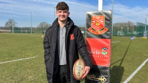 Spencer has brown hair and is wearing a grey top and black coat. He is holding a white and red rugby ball. He is standing on a green rugby pitch with a rugby post behind him, that has a padded material over it with the Keele University logo on a red background