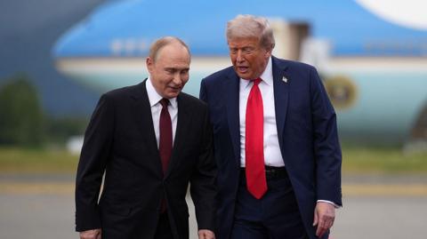 Vladimir Putin and Donald Trump, both in blue suits and red ties, talk as they walk at an airbase in Alaska