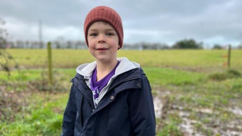 Solly smiles at the camera as he stands in front of a muddy field. He has a dark blue thick coat on and a dull pink woolen hat. 