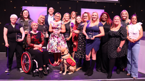 All the winners of the Make a Difference Awards standing together on a stage in fancy clothes. They are all smiling and some are holding their awards.