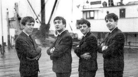 Image shows George Harrison, John Lennon, Ringo Starr and Paul McCartney in 1962, standing on the landing stage at Liverpool, with the distinctive mast of the Salvor behind them. It has three masts that point outwards on an angle. 