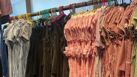 A shop rail with various school uniform dresses hanging down. They include blue, pink and green check dresses and dark tunics.