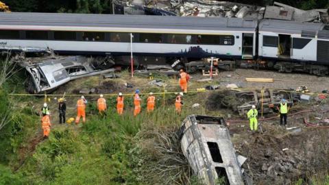 Stonehaven derailment - image of a train on tracks in woodland, and also a burned carriage down a slope, with workers in orange overalls.
