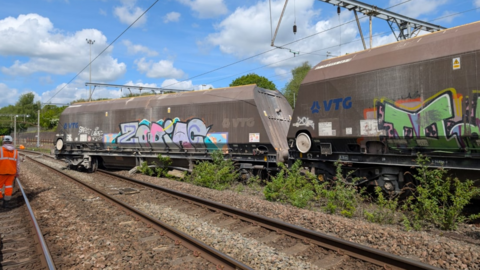 A wagon has come off a track and sits on another neighbouring line. Two wagons can be seen in the image, both what looks like a dirty brown or metal colour with graffiti on. Some rail workers stand nearby. 