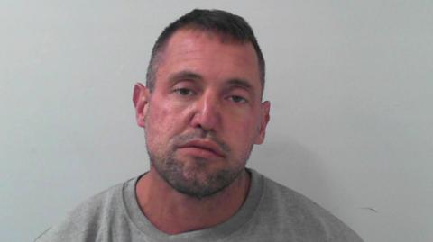 A police mugshot of a man with short dark hair and dark stubble. He has a motionless expression.