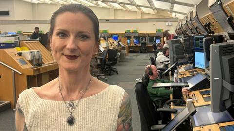 A woman smiling in a large room with banks of computers at desks. People are sat at large screens wearing headphones. She has her dark hair tied back and is wearing a white vest top and two necklaces. She has a tattoo on her shoulder.