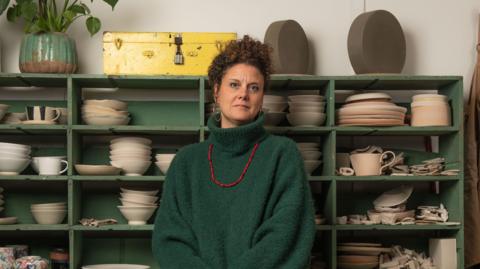 A woman in a green turtle neck jumper and red necklace sitting in front of a set of green shelves with various pieces of pottery on them