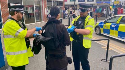Two police officers wearing black hats and high vis vests over their black uniform stand either side of the youth, whose back is turned to us. He is dressed all in black with a black rucksack that is open and being searched by the officers on the left. They stand on a pedestrianised area next to several shops and a blue and neon police car is parked next to them, by double yellow lines.