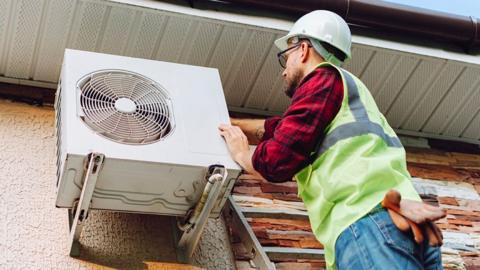 Technician in high vis vest and hardhat installs an air heat pump on the outside wall of a house