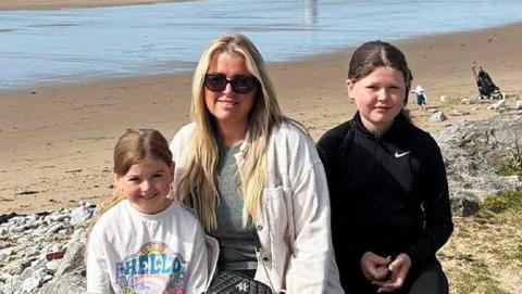 A young white girl wearing a t-shirt saying hello, her mother and her older sister sit on a rock at the edge of the beach. The mother, Lucy, has long blonde hair and wears black trousers, a plain tshirt and a white light jacket and black handbag over it. She is wearing sunglasses. Lola to her right is aged about 11, is wearing black trousers and sport top. all three are smiling at the camera. It's a sunny day. A few people sit on the beach behind them.