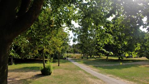A path runs through an area of mowed grass surrounded by large deciduous trees. The photograph has been taken in the summer and parts of the short grass is parched.