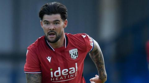 Alex Mowatt in action for West Bromwich Albion. He is wearing a red away shirt. It is short sleeved and shows his heavily tattooed armss