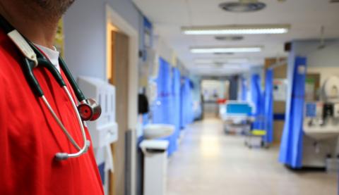 A doctor wearing a stethoscope stands in front of a hospital ward with beds and hospital screens 