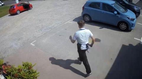 Young man wearing a white shirt and apron, running through a car park towards a red car