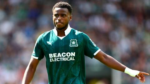 Plymouth Argyle's Bali Mumba looks ahead and pokes his tongue out as he points with his left arm out to the side. he has strapping round his wrist and wears a dark green patterned shirt with white trim.