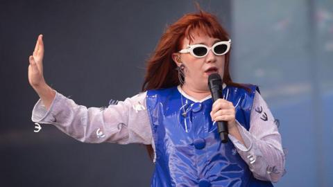 A woman with red hair is singing on a stage. She has a microphone in her left hand and is holding it to her mouth. Her other hand is raised at her side. She is wearing white sunglasses and a blue dress with white sleeves. 