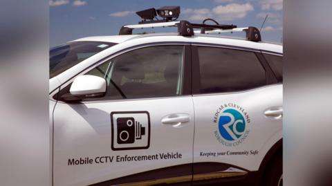 A white car with two black cameras mounted on top. Stickers printed on the side read: "Mobile CCTV Enforcement Vehicle" and "Keeping you Community Safe". The Redcar and Cleveland Borough Council logo is also printed on the car door, as is an icon depicting a camera.