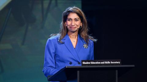 Suella Braverman is standing in front of a backdrop and in front of a black podium. There is a black name sign with white writing that reads "shadow education and skills secretary". She has brown hair and is wearing a blue suit jacket with black buttons. She has a large silver brooch on the jacket and a nude coloured microphone wire near her mouth