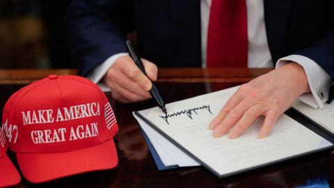 US President Donald Trump signs an executive order in the Oval Office at the White House in Washington, DC on 23 April 2025. A red baseball hat embroidered with "Make America Great Again" in white thread sits to the left of the document, which shows Trump's distinctive signature