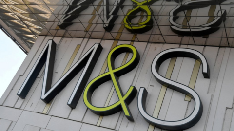 M&S sign on building with reflection in roof. White M and S lettering and the & sign is yellow.