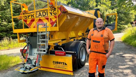 Road worker in orange high-visibility clothing standing beside a yellow gritting truck marked Spreading on a rural roadside with trees in the background.