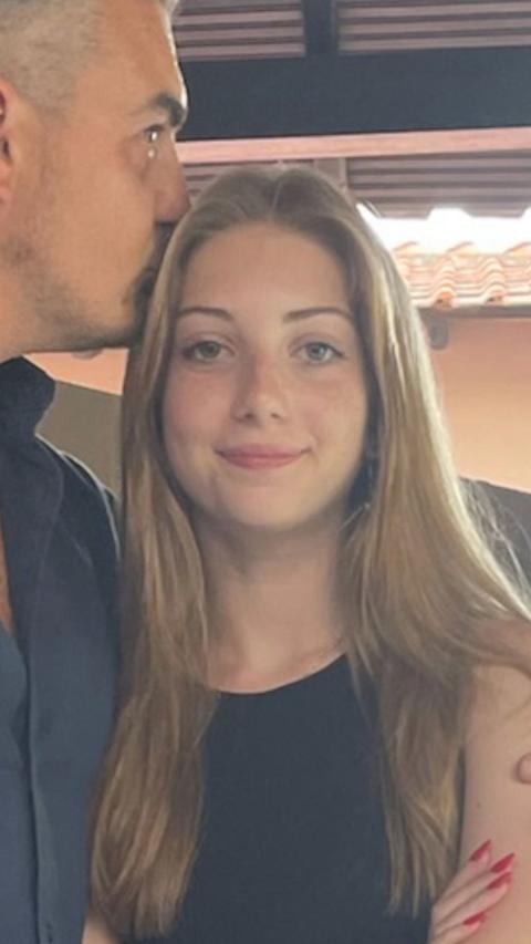 A young woman with long brown hair wearing a black top stands next to a man, who is kissing her head