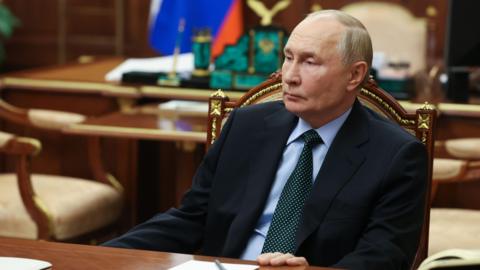 Vladimir Putin sits at a wooden table, wearing a black suit jacket, a blue shirt and a spotted green tie.