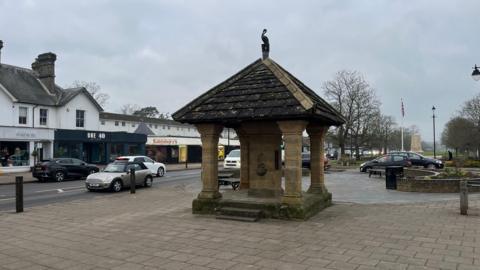 An image of the centre of Cranleigh, including a former drinking fountain, which has become a well known monument in the village. Cars drive past on the high street. Shops, including Sainsbury's, are in the background. Also in the background is the local war memorial.