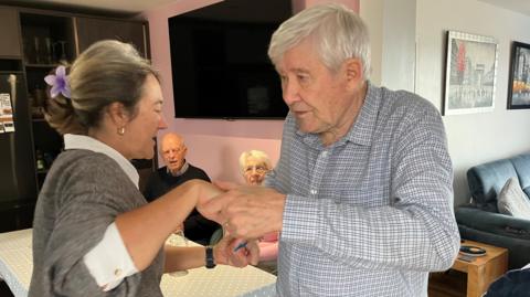 A lady is holding hands with an elderly gentlemen with two other people at a table looking on.
