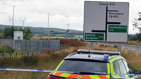 a police car is parked behind a cordon and in front of a road sign showing directions to Claudy, Dungiven, Belfast, Londonderry and Dungiven. In the backgorund is a second police car and a number of wind turbines can be seen atop hills in the distance