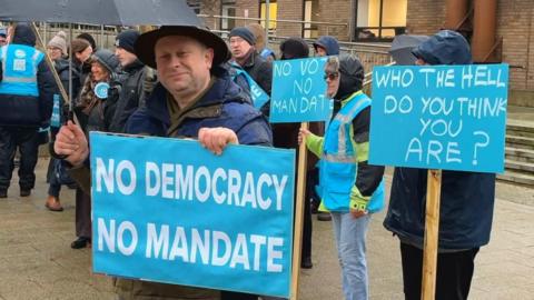 A group of people, mostly men, stand in the drizzle holding blue-coloured placards, that read NO DEMOCRACY NO MANDATE and WHO THE HELL DO YOU THINK YOU ARE?. A man stands in the foreground, holding an umbrella, looking towards the camera