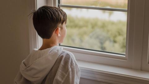 Young boy looking out of a window, wearing a white hoodie. The hood is hanging down his back. The boy is viewed from the back of his head, with the window in view.
