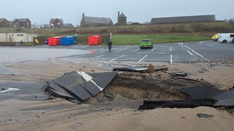The sinkhole in a tarmac car park. The hole is about as large as three parking spaces. The area surrounding the hole has broken sections of tarmac and sand around it. One side of the car park is covered in water. A handful of cars are parked some distance away from the hole.