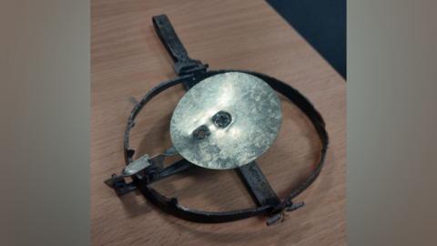 A gin trap. It comprises of a silver metal disk with a black metal ring around it. It is placed on a wooden table.