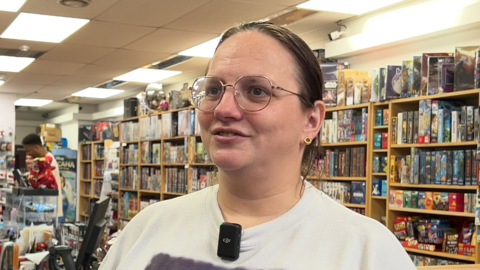 Becky Ottery is standing in her games shop, which has many shelves of games behind her. She is wearing a white T-shirt and has long, brown hair, which is tied up.