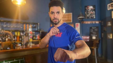 A man with a goatee beard holds his fist up to the camera in a fight pose