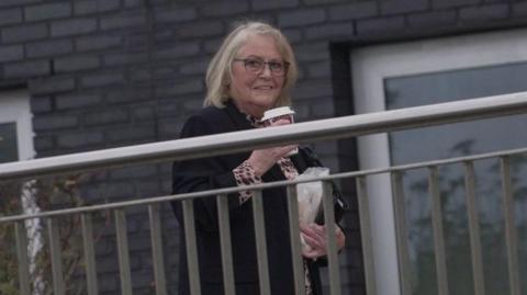Sharman behind a metal silver fence walking into court. She is smiling and looking at the camera. She carries a takeaway coffee and a bag in her other hand.