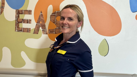 Amy Carberry in nurse uniform smiling at the camera with her hands in her pocket. She has long blonde hair tied back. She is stood in front of a wall with colourful patterns on.