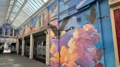 The mural painted on the wall inside Lowther Arcade. A large section of wall shows two geese flying through a blue sky with their wings outstretched. behind them are clouds which are coloured pink, orange and purple as if catching the light from the sun. The mural stretches out above shop fronts inside the arcade.