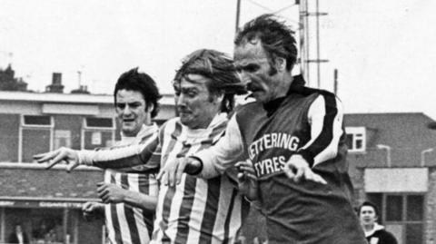 A black and white image of Derek Dougan wearing Kettering Town FC's infamous sponsored kit. It has "Kettering Tyres" printed on the front. Dougan is jostling for the ball with two opposition players.