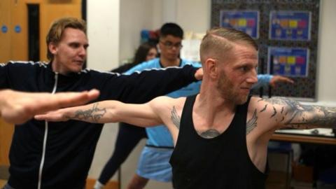 Adam Townsend is striking a yoga pose, where his arms are outstretched. He is heavily tattooed, with a brown beard. He is wearing a black vest. Other members are doing the same pose in the background. 